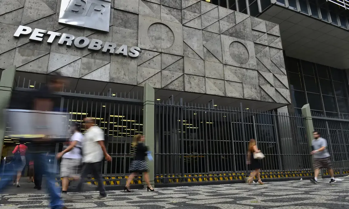 Pessoas caminham em frente ao edifício sede da Petrobras, com letreiro da empresa em destaque na fachada de concreto.