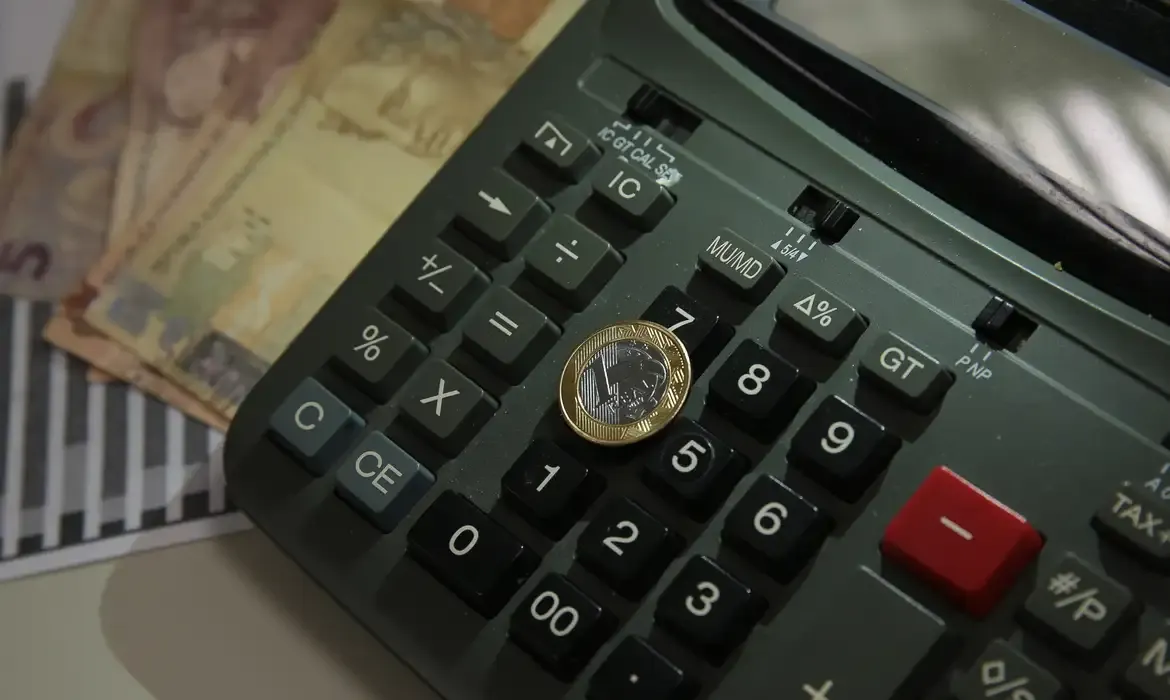 Moeda de um real sobre teclado de calculadora preta, com cédulas de dinheiro ao fundo.