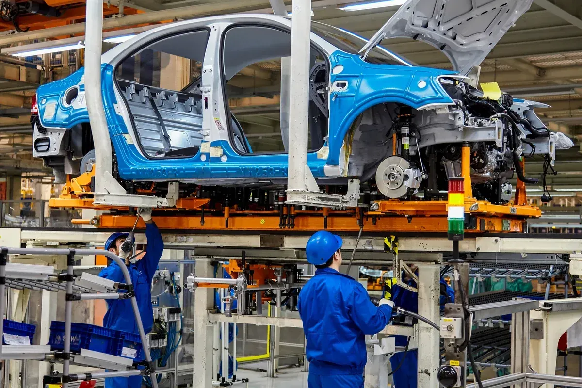 Trabalhadores com uniformes e capacetes azuis operam em linha de montagem de carcaça de veículo em fábrica automotiva.