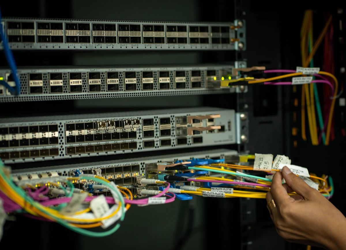 Mão humana ajustando cabos de fibra óptica coloridos conectados a servidores em um rack de data center.