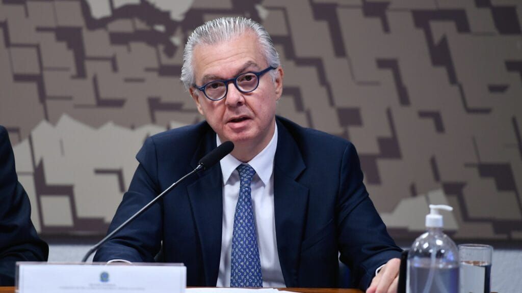 O embaixador Rodrigo de Azeredo Santos, de óculos e terno azul, fala ao microfone durante uma mesa de reunião.