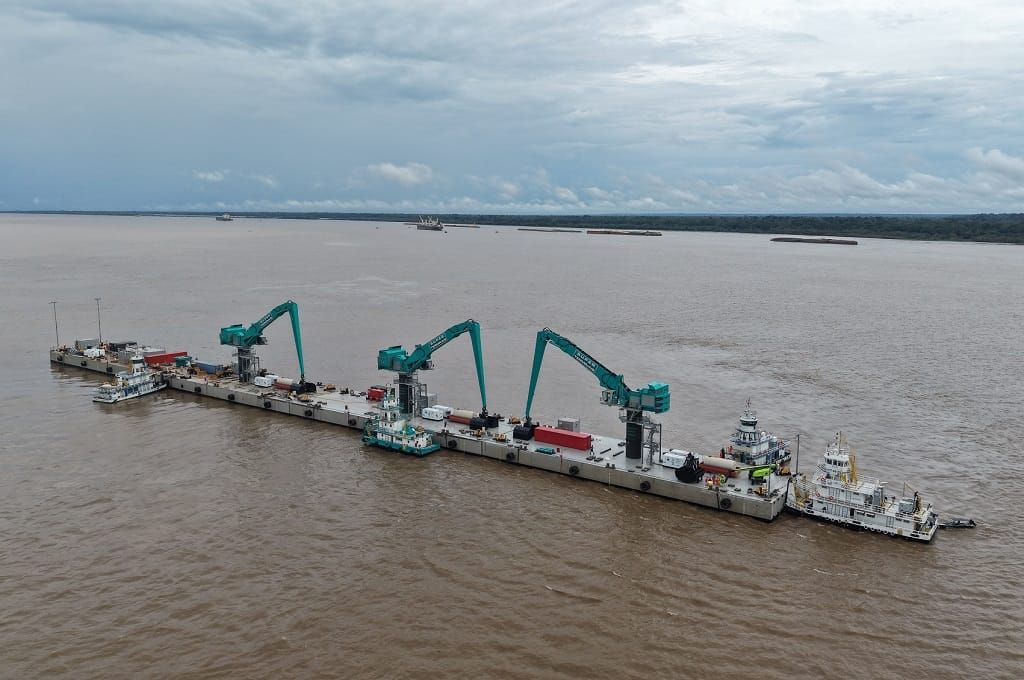 Píer flutuante com guindastes verdes operando no meio de um rio largo sob céu nublado.