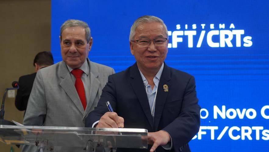 Gilberto Takao Sakamoto, presidente do CFT, assina documento em púlpito transparente diante de painel azul do Sistema CFT/CRTs.