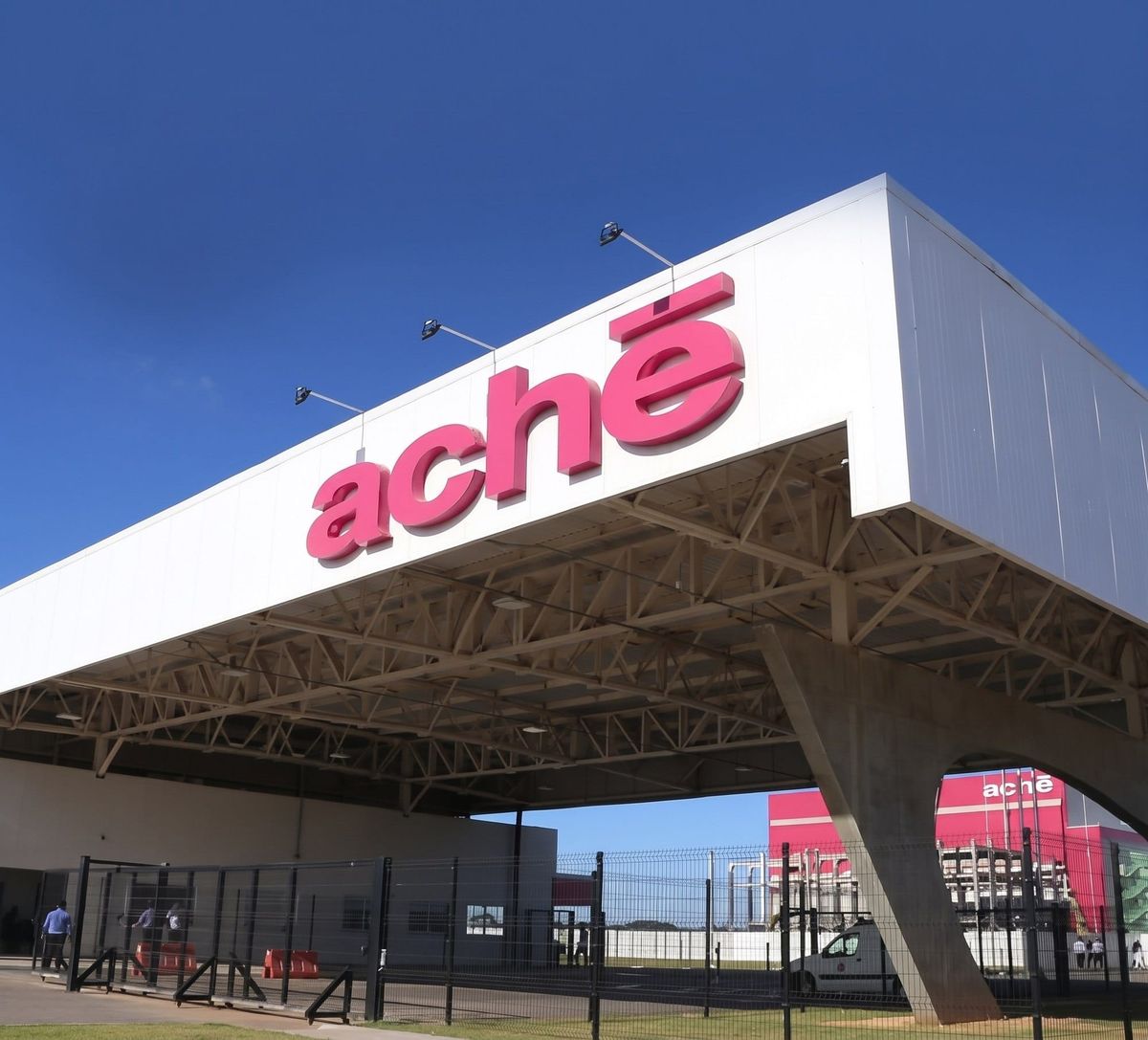 Logotipo rosa do Aché em destaque na cobertura branca da portaria de uma fábrica, com galpões ao fundo sob céu azul.