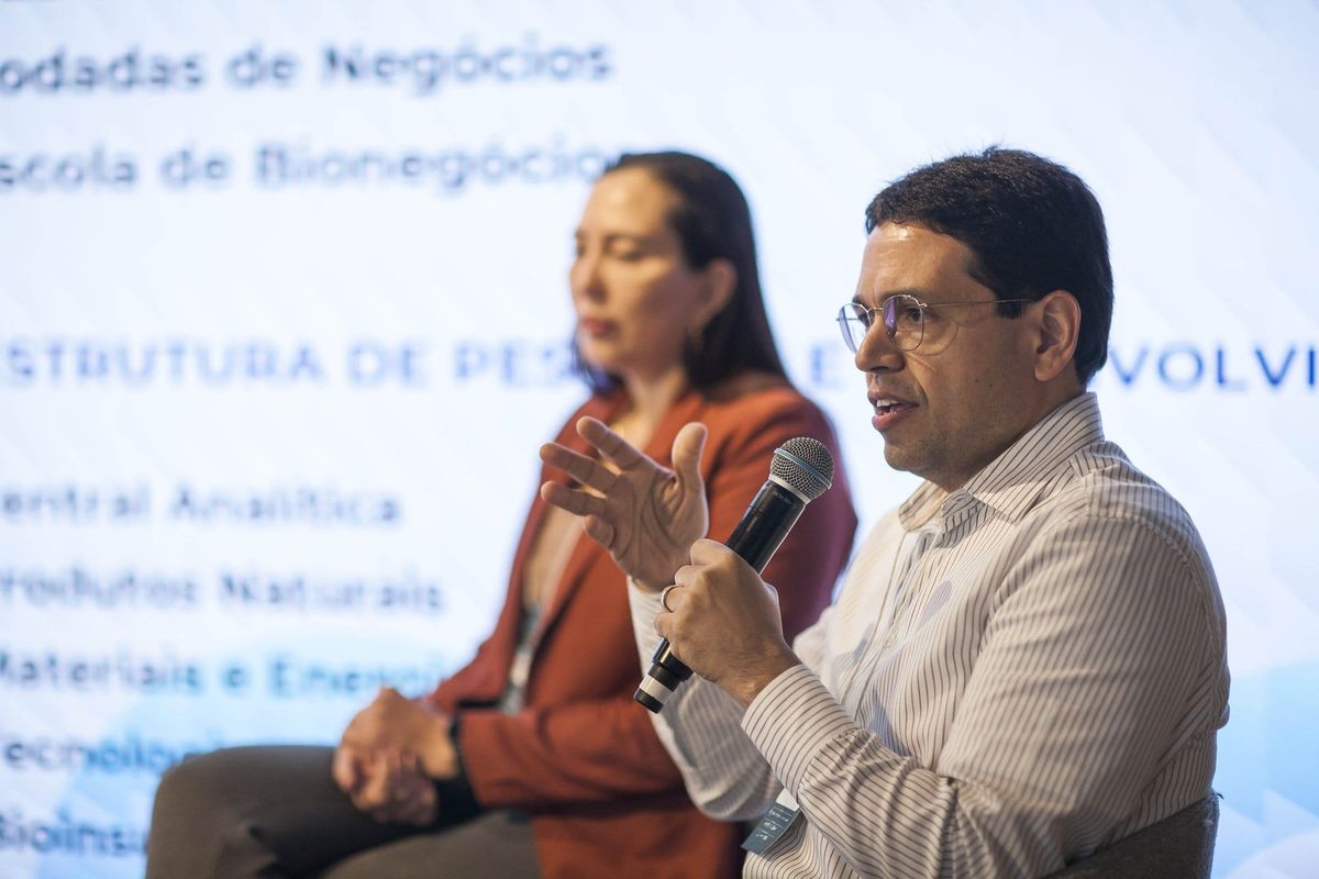Carlos Henrique Carvalho fala ao microfone no palco, com Andréia Freitas Vieira ao fundo e slide da estrutura do CBA