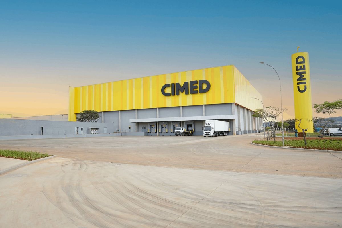Galpão amarelo da Cimed com docas, caminhões e torre cilíndrica com o nome da empresa em Pouso Alegre