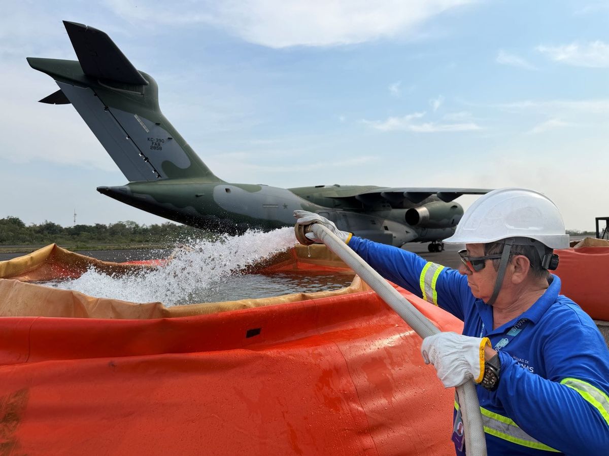 Funcionário da Águas de Manaus direciona mangueira de água para tanque laranja em frente a uma aeronave militar KC-390.