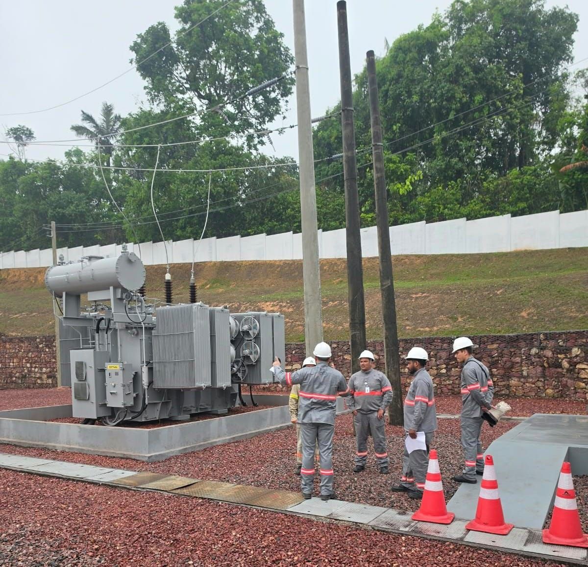 Trabalhadores com uniformes cinza e capacetes brancos analisam transformador de alta potência em subestação de energia.