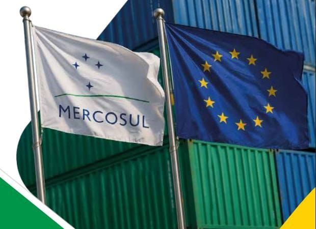 Bandeiras do Mercosul e da União Europeia hasteadas em frente a contêineres de carga empilhados.