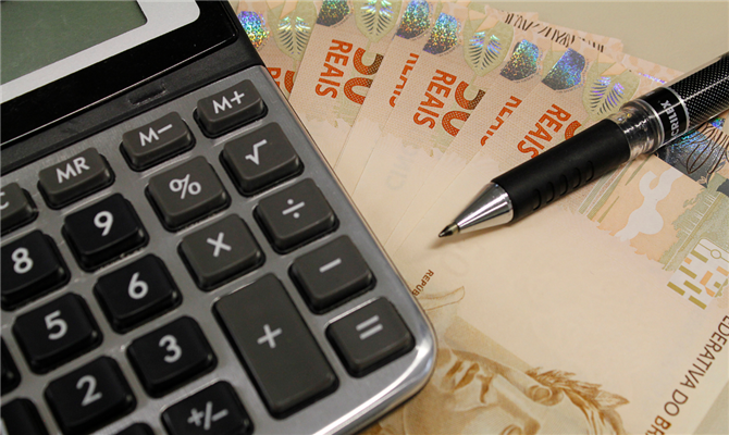 Calculadora sobre notas de 50 reais e uma caneta, representando o cálculo de dívidas e inadimplência de empresas.