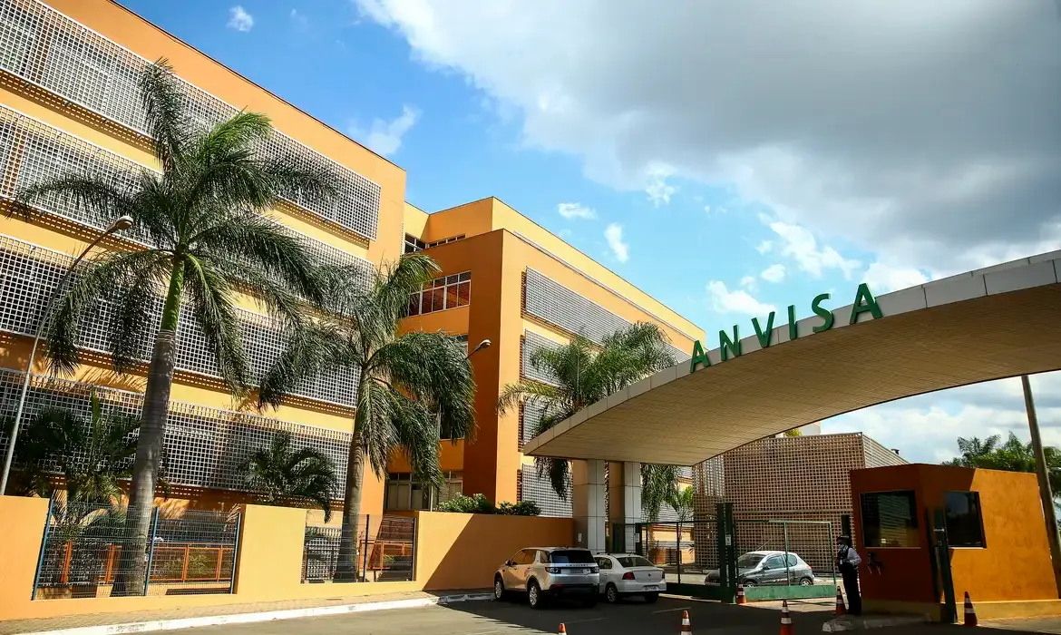 Fachada do edifício da Anvisa com palmeiras à frente e carros estacionados na entrada sob céu azul.