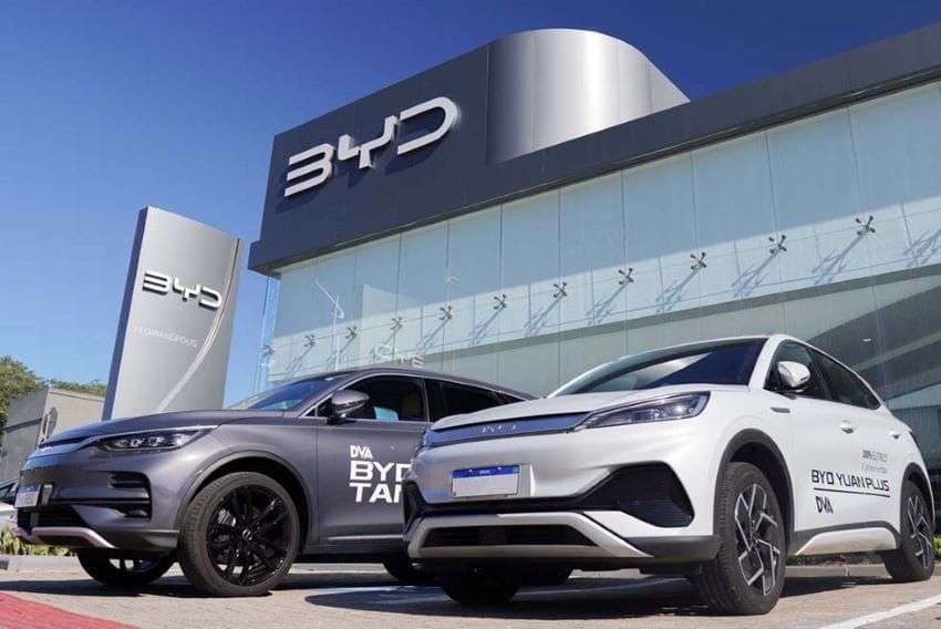 Dois veículos da BYD, um cinza e um branco modelo Yuan Plus, estacionados em frente a uma concessionária da marca.