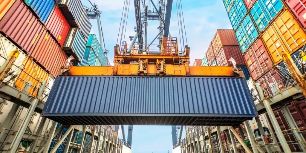 Guindaste portuário içando um contêiner azul em meio a pilhas de contêineres coloridos em um terminal de carga.