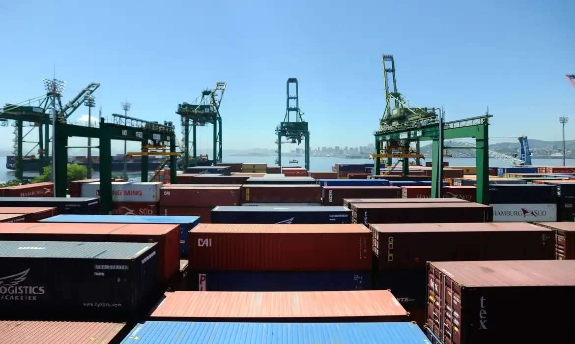 Contêineres coloridos empilhados em terminal portuário com guindastes verdes ao fundo sob céu azul.