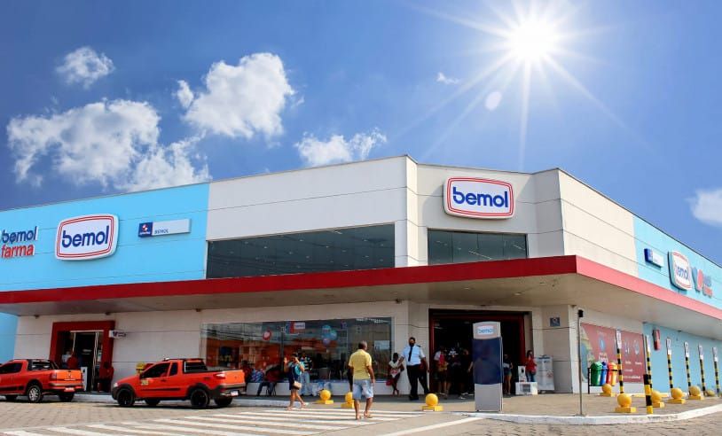 Fachada de uma unidade da Bemol com as marcas Bemol Farma e Bemol em destaque sob céu azul e sol forte.