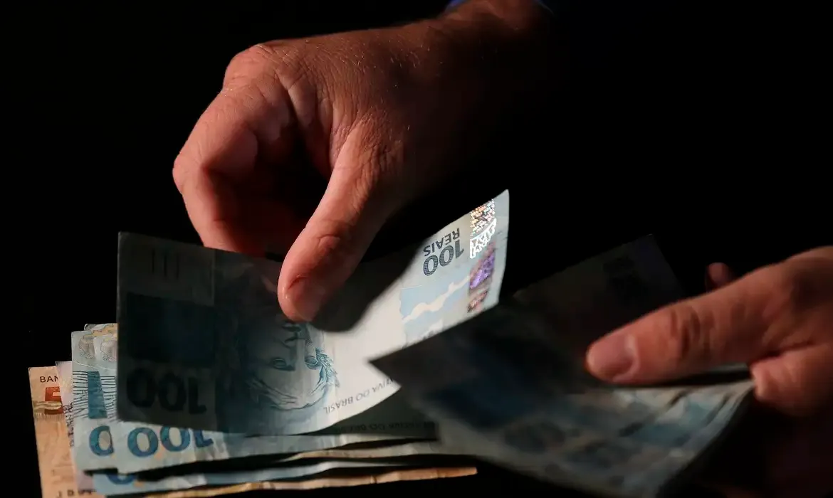 Mãos de uma pessoa contando diversas cédulas de cem reais em um ambiente com fundo escuro.