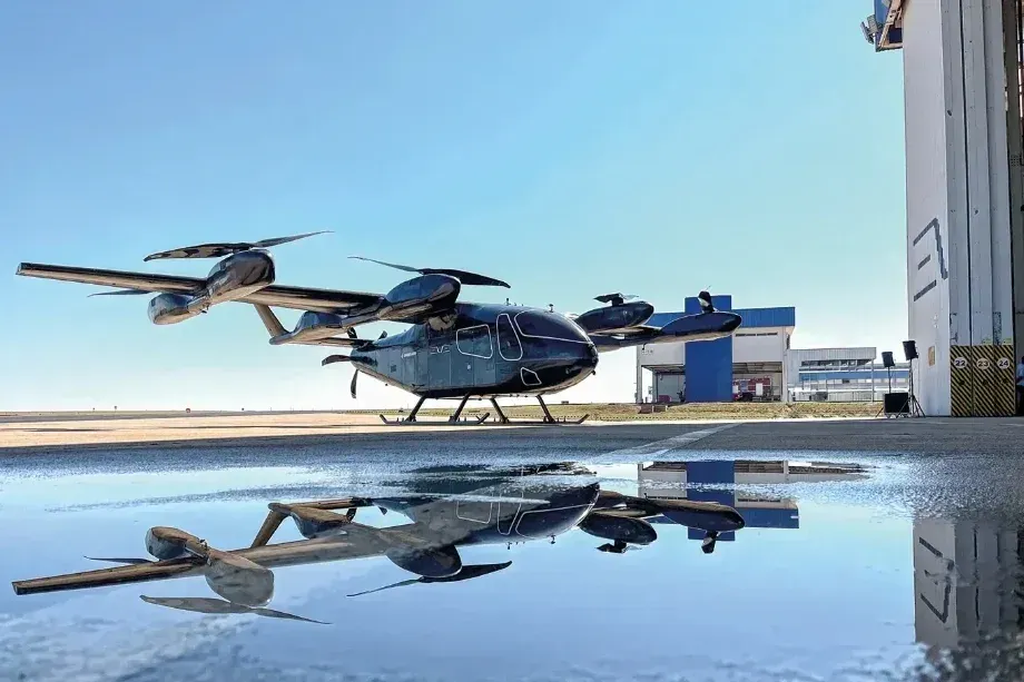 Aeronave elétrica preta eVTOL da Eve Air Mobility estacionada em pátio de hangar com reflexo em poça d'água.