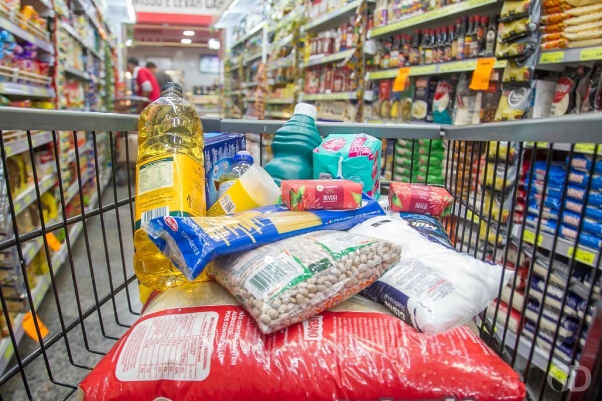 Carrinho de supermercado abastecido com itens como óleo, feijão, arroz e macarrão em um corredor de loja.