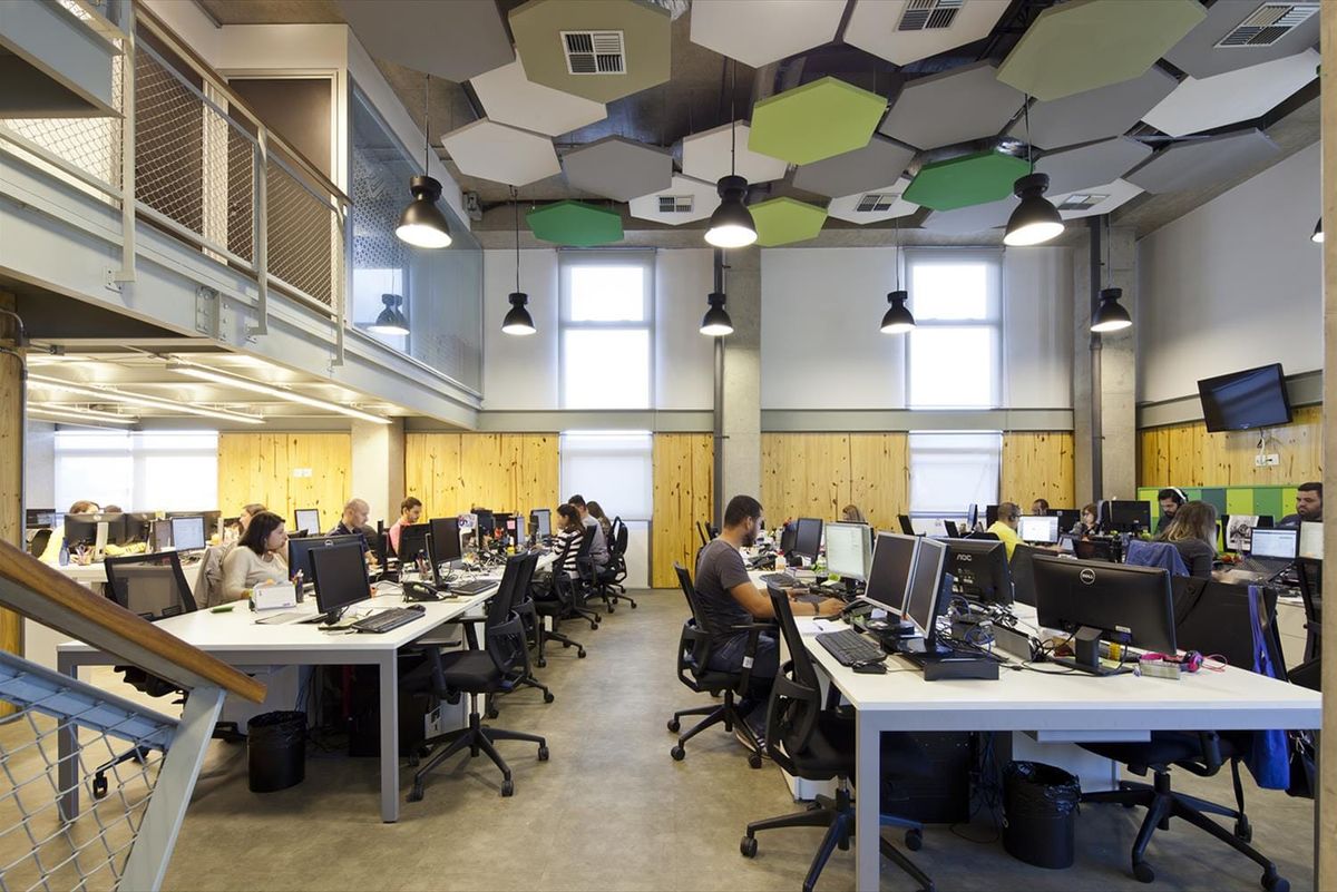 Colaboradores trabalhando em computadores em um escritório moderno com pé-direito alto e decoração industrial.