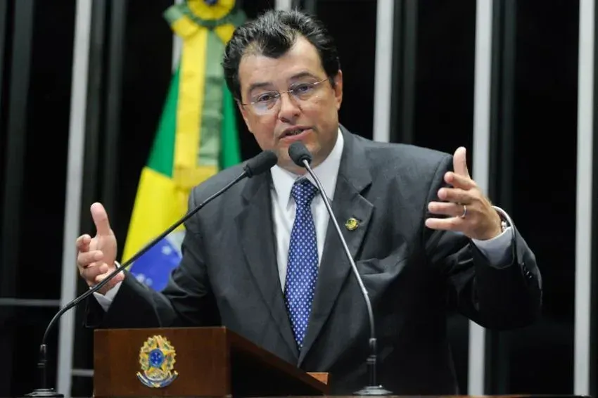 O senador Eduardo Braga discursa em um púlpito com microfones, vestindo terno escuro e gravata azul, com a bandeira do Brasil ao fundo.