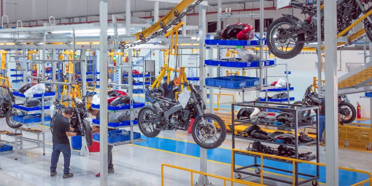 Linha de montagem da fábrica da Bajaj em Manaus, com motocicletas suspensas em suportes amarelos e funcionários trabalhando.