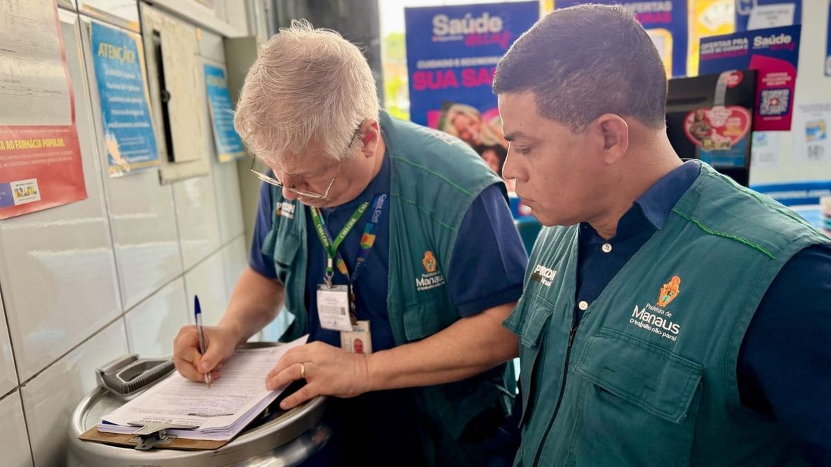 Dois fiscais do Procon Manaus, usando coletes verdes da prefeitura, preenchem formulário durante fiscalização em farmácia.