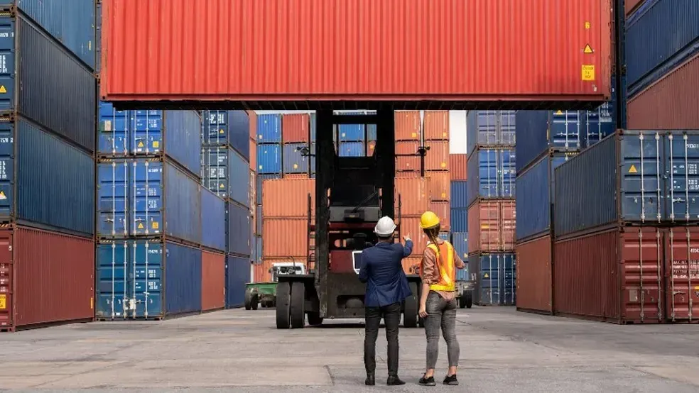 Dois trabalhadores de capacete observam empilhadeira movimentando contêiner vermelho em terminal portuário.