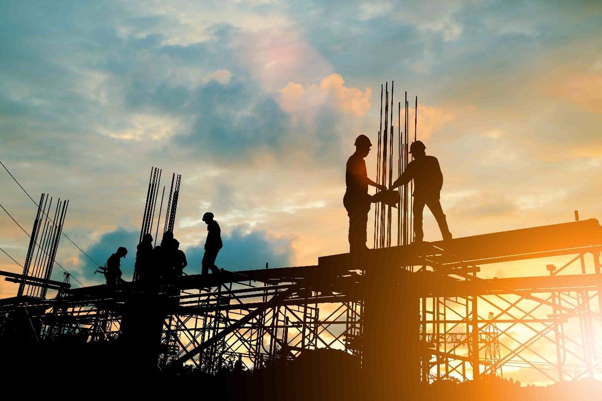 Silhueta de trabalhadores em canteiro de obras de infraestrutura sob o pôr do sol, com vigas de aço e andaimes expostos.