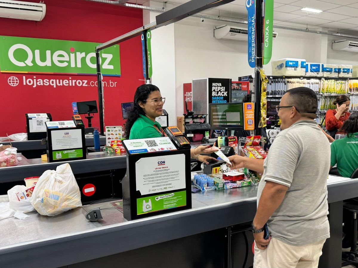 Operadora de caixa atende cliente em unidade das Lojas Queiroz em Manaus, com logomarca da rede ao fundo.