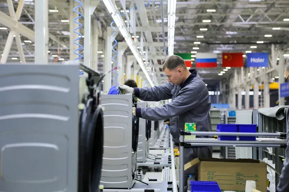 Trabalhador em linha de montagem de máquinas de lavar da Haier, com bandeiras da China e Rússia ao fundo.