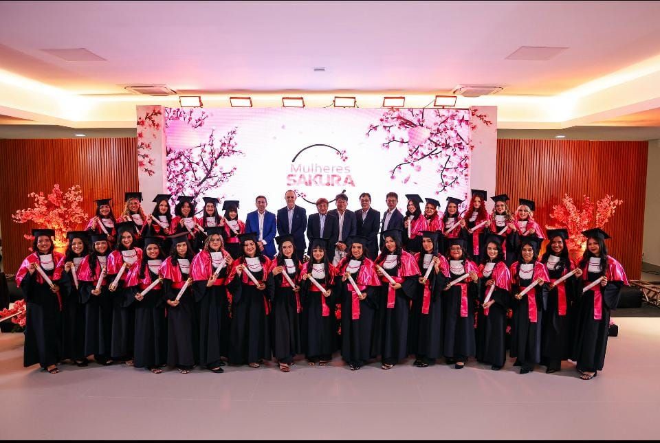 Grupo de mulheres formandas vestindo becas pretas e faixas rosas posam com diplomas no palco do Projeto Mulheres Sakura.