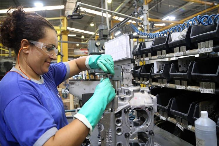 Trabalhadora com luvas verdes e óculos de proteção manipula peça metálica em linha de montagem industrial.
