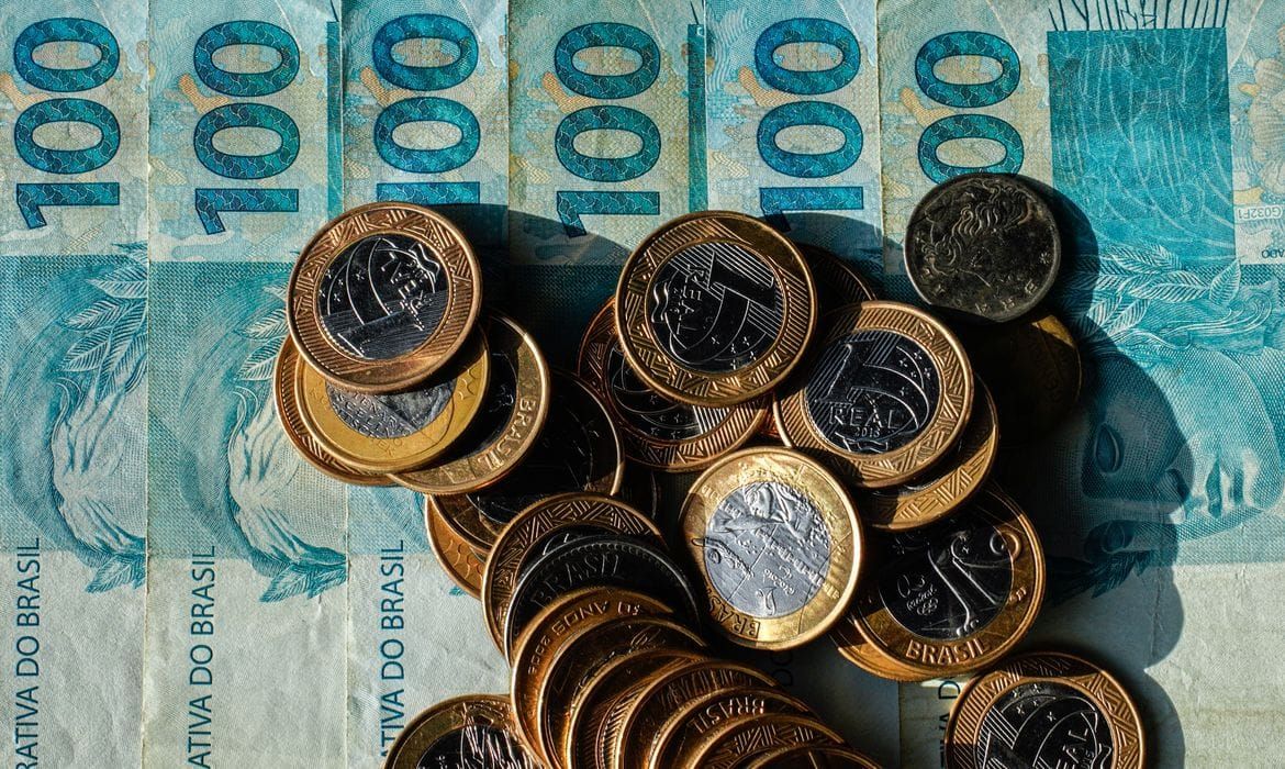 Cédulas de cem reais dispostas verticalmente com diversas moedas de um real espalhadas por cima.