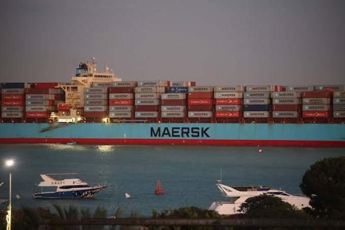 Navio porta-contêineres azul da Maersk carregado navegando em um rio ao entardecer, com lanchas menores em primeiro plano.