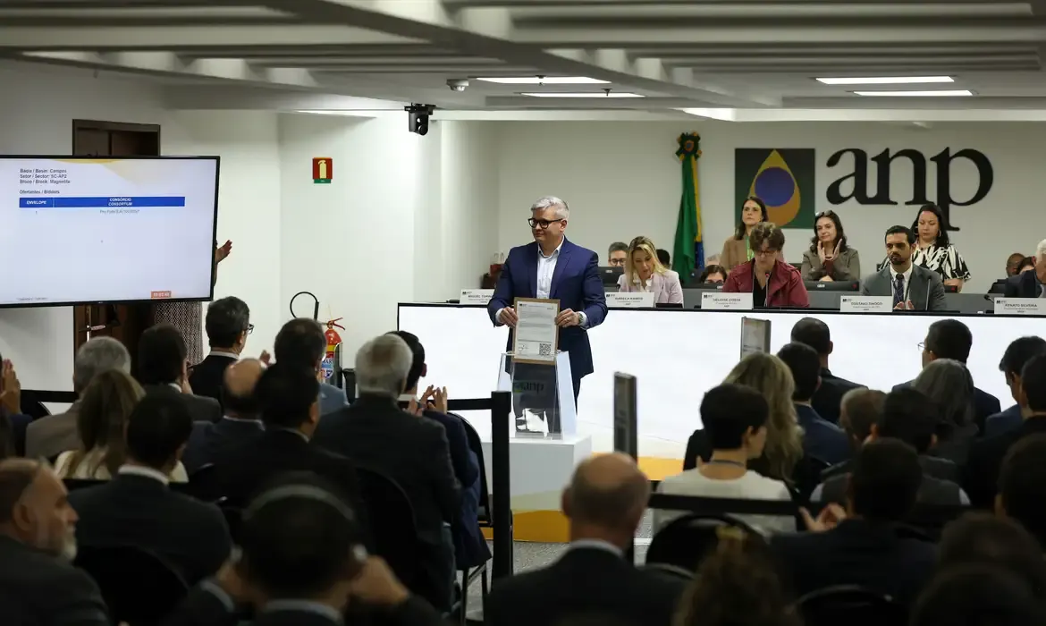 Homem de terno azul segura documento diante de plateia e telão durante leilão na sede da ANP, no Rio de Janeiro.