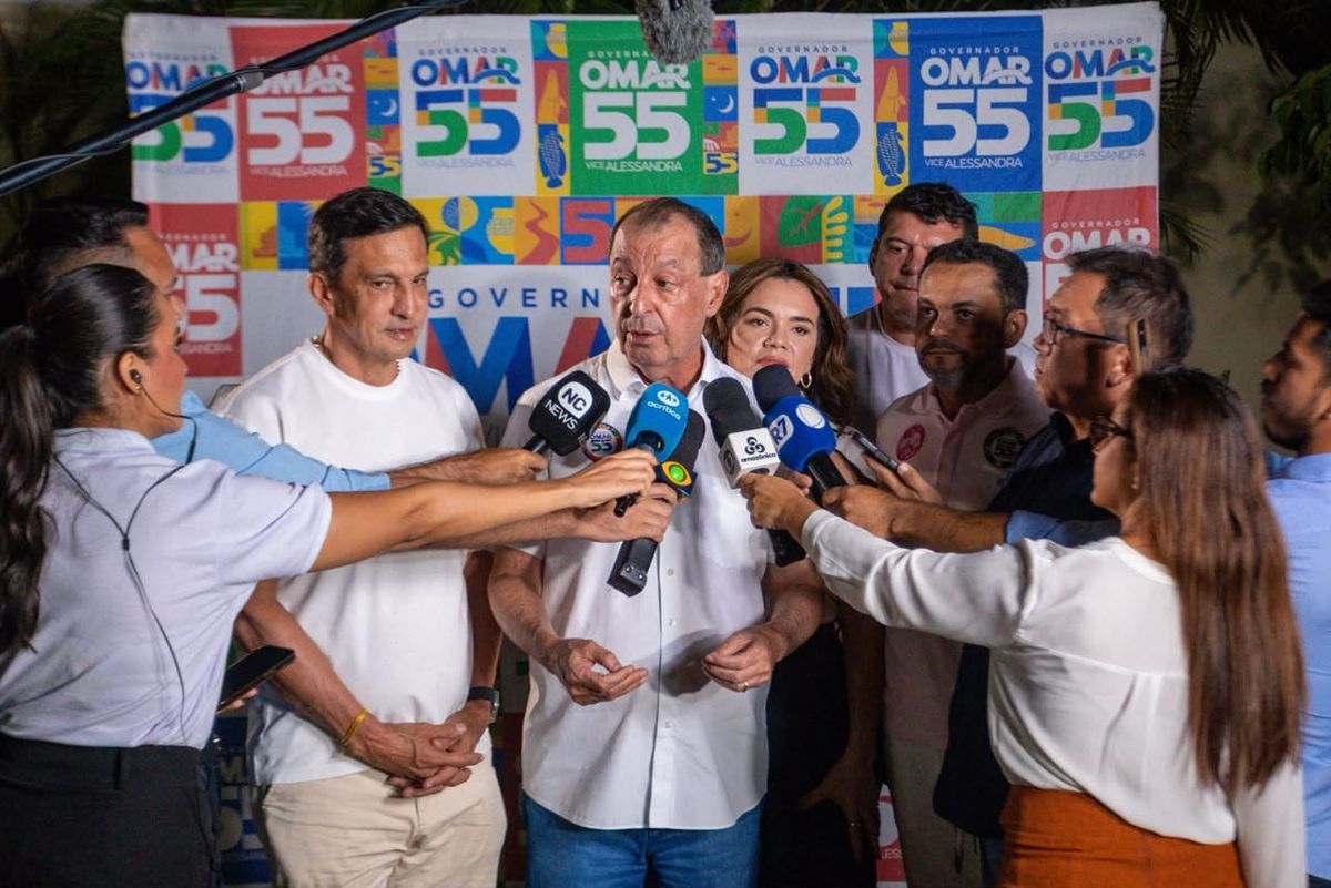 O candidato Omar Aziz concede entrevista coletiva cercado por jornalistas e microfones em frente a painéis de sua campanha.
