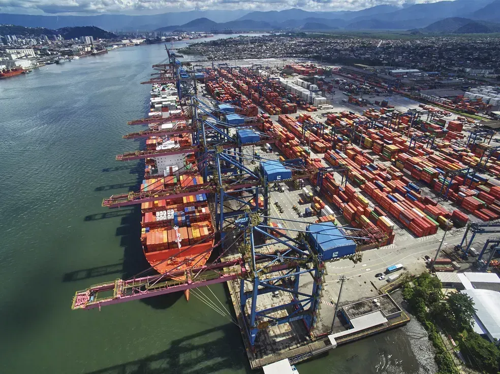 Vista aérea de terminal portuário com navio cargueiro sendo operado por guindastes e pátio repleto de contêineres coloridos.