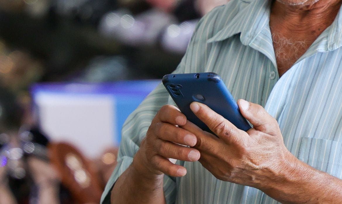 Homem segura smartphone azul com as duas mãos, em plano fechado, sugerindo o uso de aplicativos.