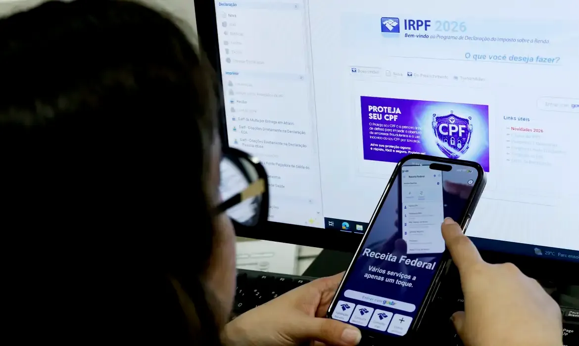 Pessoa segura celular com aplicativo da Receita Federal aberto em frente a monitor com o programa do IRPF 2026.