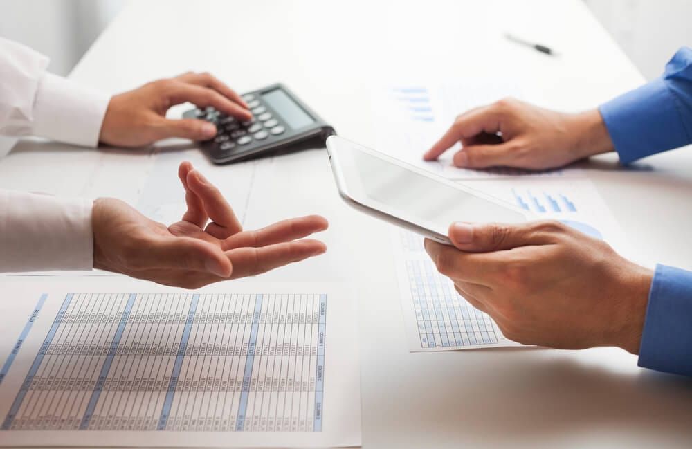 Duas pessoas em uma mesa com calculadora, tablet e papéis com tabelas financeiras, simulando uma negociação de dívidas.
