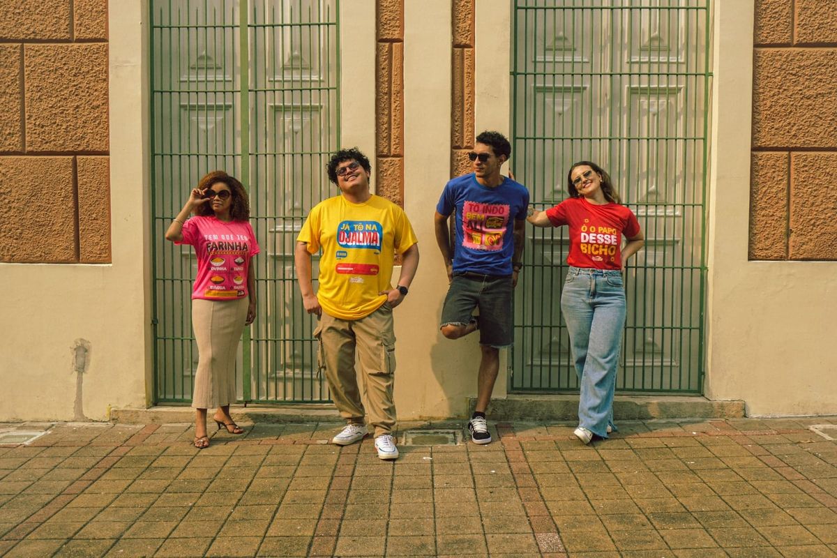 Quatro pessoas posam em frente a um portão verde vestindo camisetas coloridas da coleção Manaus Raiz.