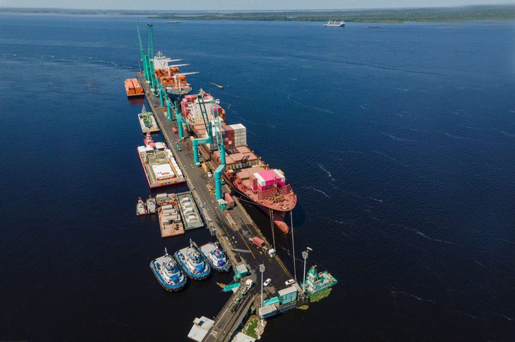 Vista aérea do Super Terminais em Manaus com navio de carga atracado, contêineres coloridos e guindastes operando no rio.