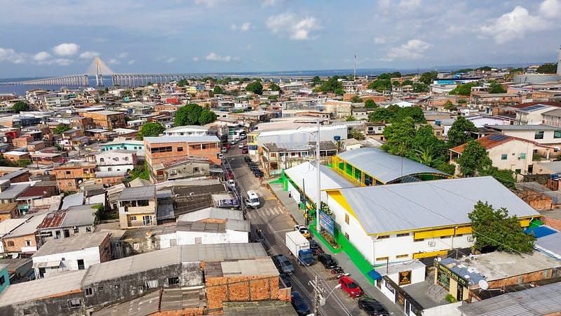 Vista aérea de área urbana densa em Manaus com casas, prédios baixos e a Ponte Rio Negro ao fundo sob céu com nuvens.