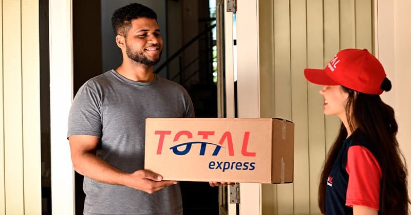 Entregadora da Total Express entrega uma caixa de papelão com o logotipo da empresa para um cliente sorridente.