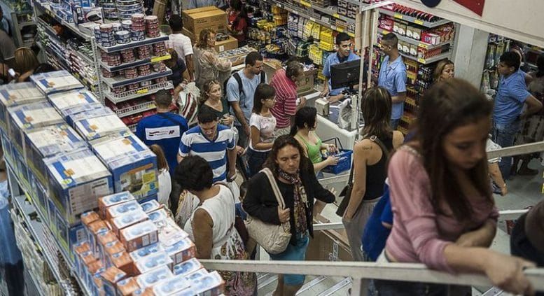 Consumidores circulam em corredor de loja de departamentos com prateleiras cheias de produtos e escada de acesso.