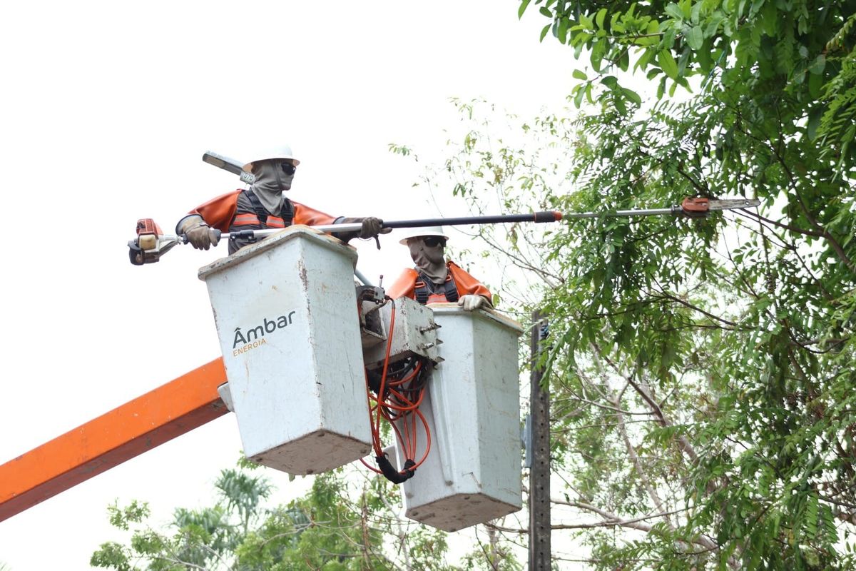 Dois técnicos da Âmbar Energia em cestos elevados realizam a poda de galhos de árvore próximos à rede elétrica.