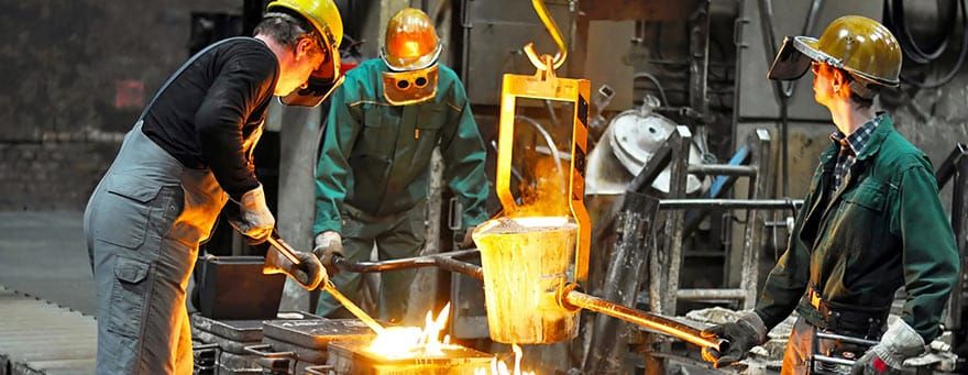 Três metalúrgicos com capacetes e roupas de proteção operando caldeira com metal fundido em uma fundição.