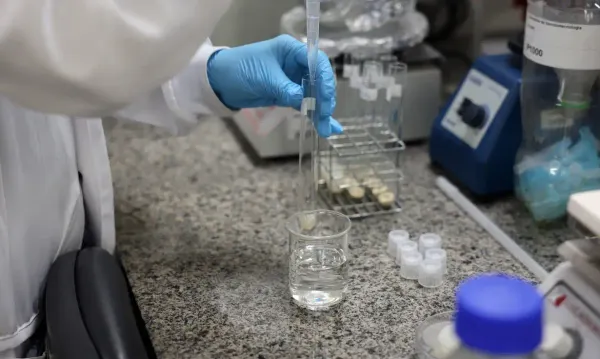 Homem de jaleco branco e óculos segurando um frasco com líquido, em ambiente de laboratório