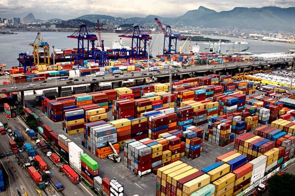 Contêineres empilhados no terminal do Porto do Rio de Janeiro, com guindastes e navio de carga ao fundo