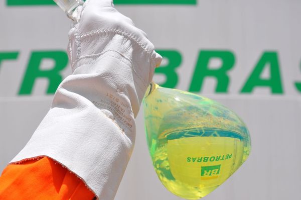 Mão com luva branca segurando um frasco de vidro com combustível amarelado em frente ao logotipo da Petrobras.